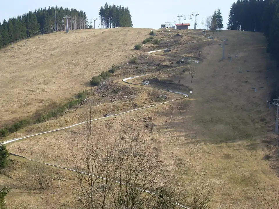 Rodelbahn in engen Serpentinen auf einem Hang mit noch braunem Gras, oben der Lift