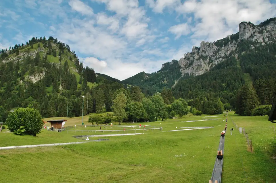 Lange Rodelrinne mit Lift in einem grünen Tal vor den Felsen der Ammergauer Alpen
