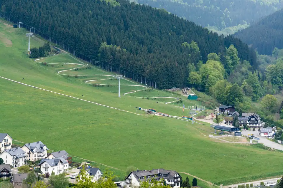 Grüner Hang mit einer Rodelbahn in vielen Serpentinen, daneben ein Lift und am Fuß die Häuser von Willingen